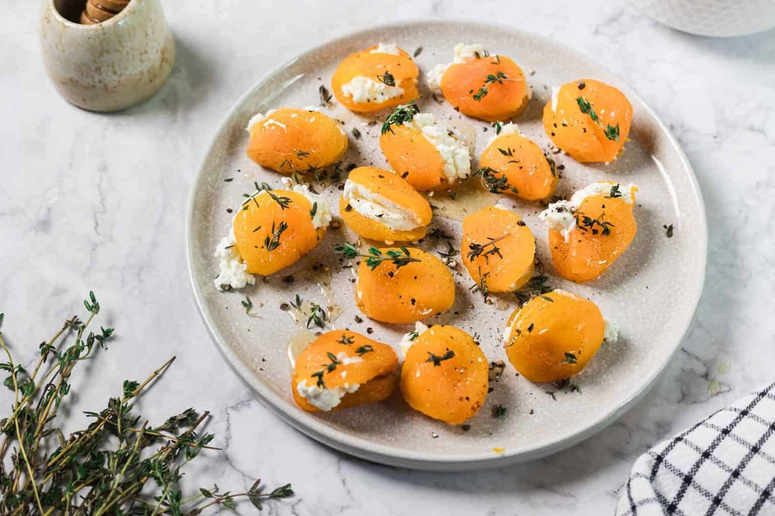 A plate of halved apricots stuffed with cheese and garnished with fresh herbs on a marble surface.