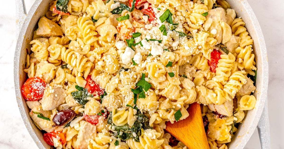 Rotini pasta with chicken, tomatoes, spinach, olives, and feta cheese in a creamy sauce, garnished with parsley.