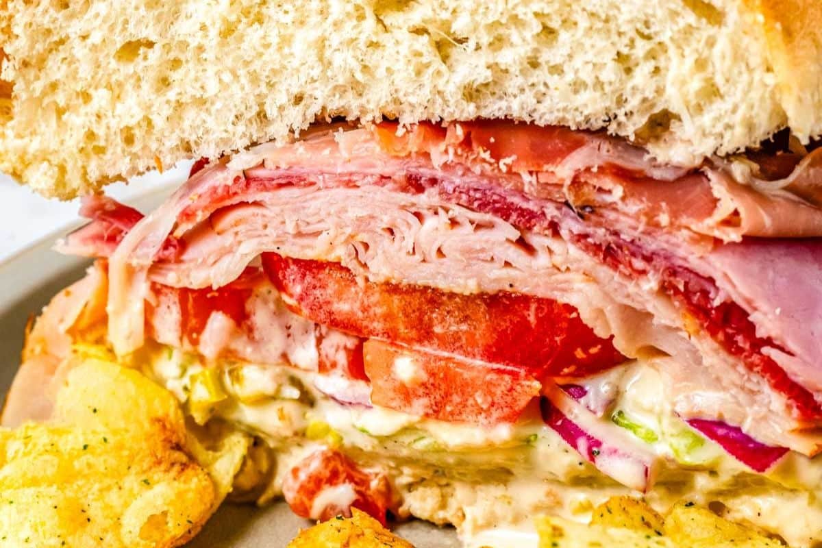 Close-up of a deli sandwich with ham, tomato, onions, lettuce, and sauce, served with potato chips.