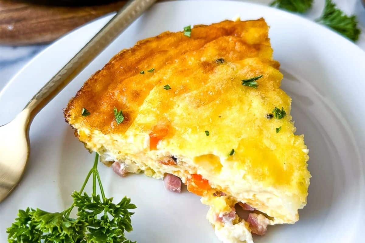 A slice of ham and vegetable quiche on a white plate, garnished with fresh parsley.