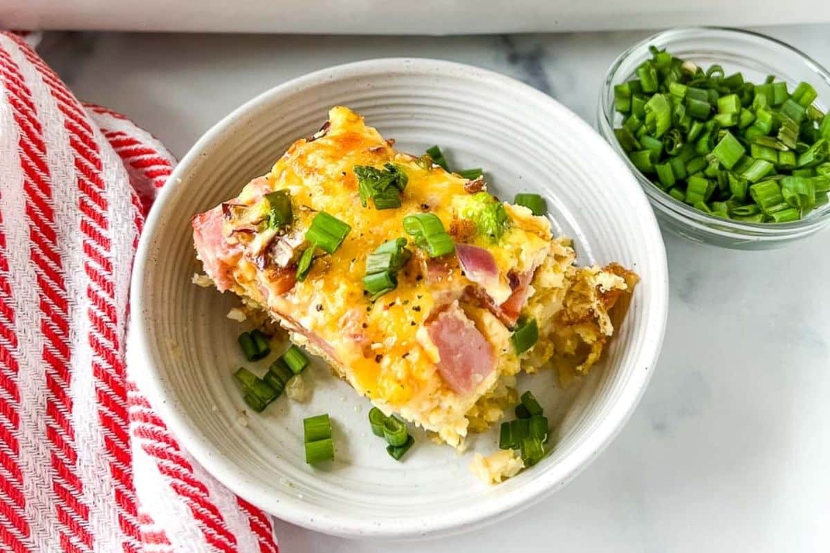 A slice of cheesy ham casserole topped with chopped green onions in a bowl, next to a dish of green onions.