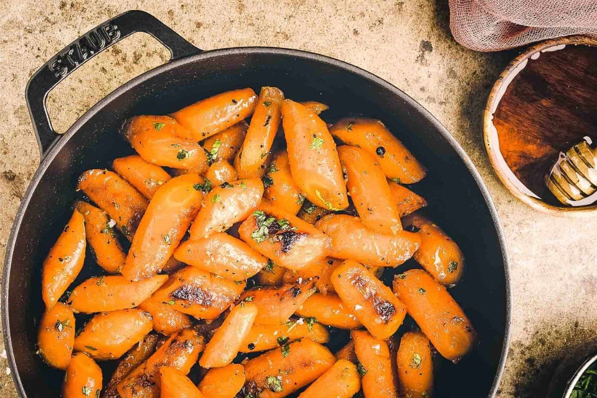 Brown cast iron pan filled with glazed roasted carrots, garnished with herbs, on a rustic surface.