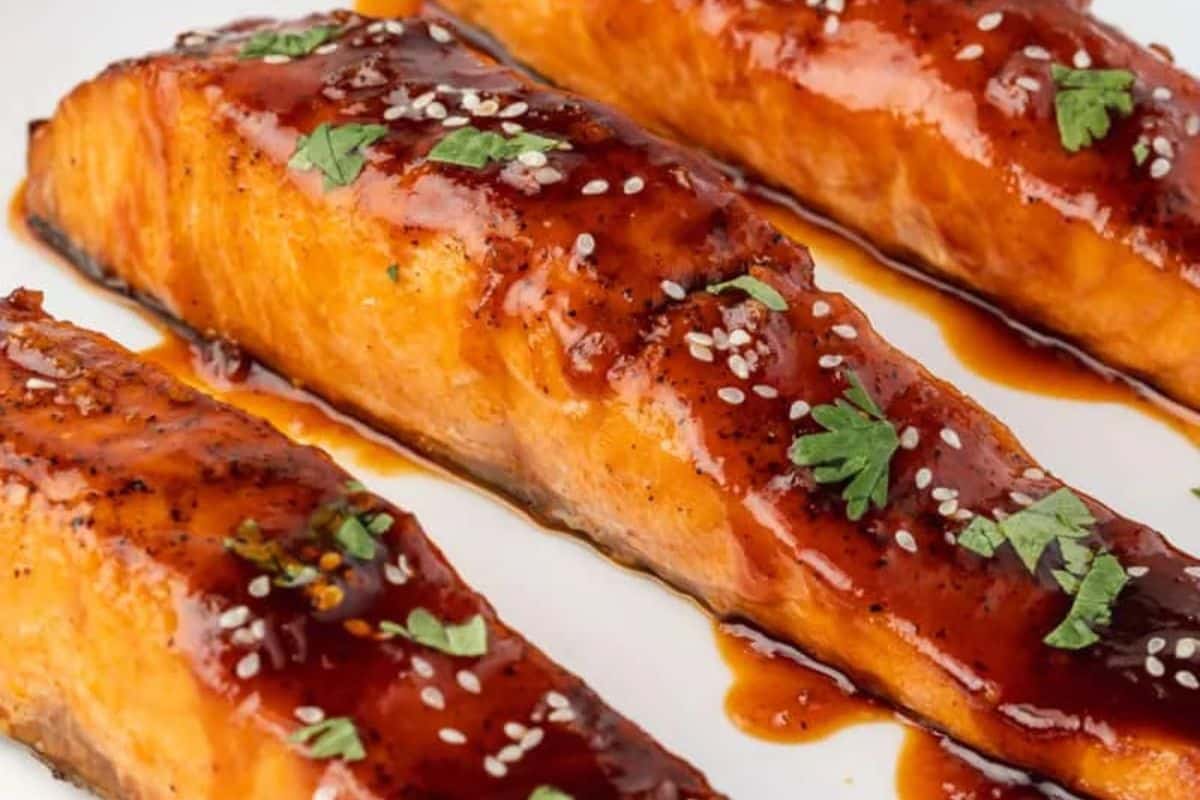 Three glazed salmon fillets topped with sesame seeds and chopped herbs on a white plate.