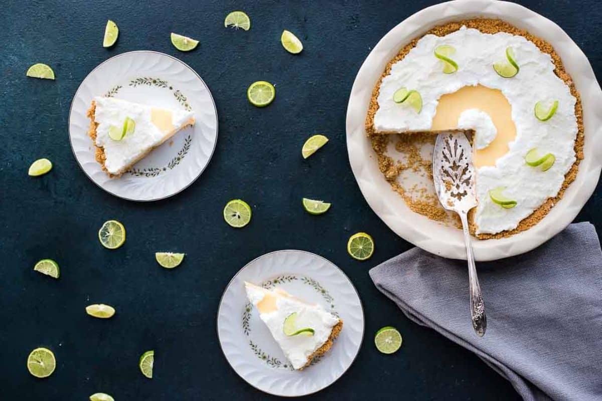 Key lime pie with whipped topping, sliced and served on plates, surrounded by lime wedges on a dark table.
