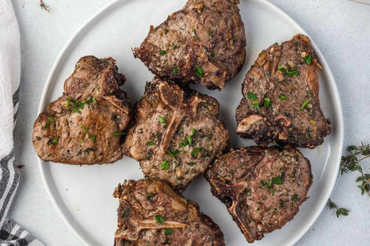 Six cooked lamb chops garnished with herbs, arranged on a white plate with a striped napkin nearby.
