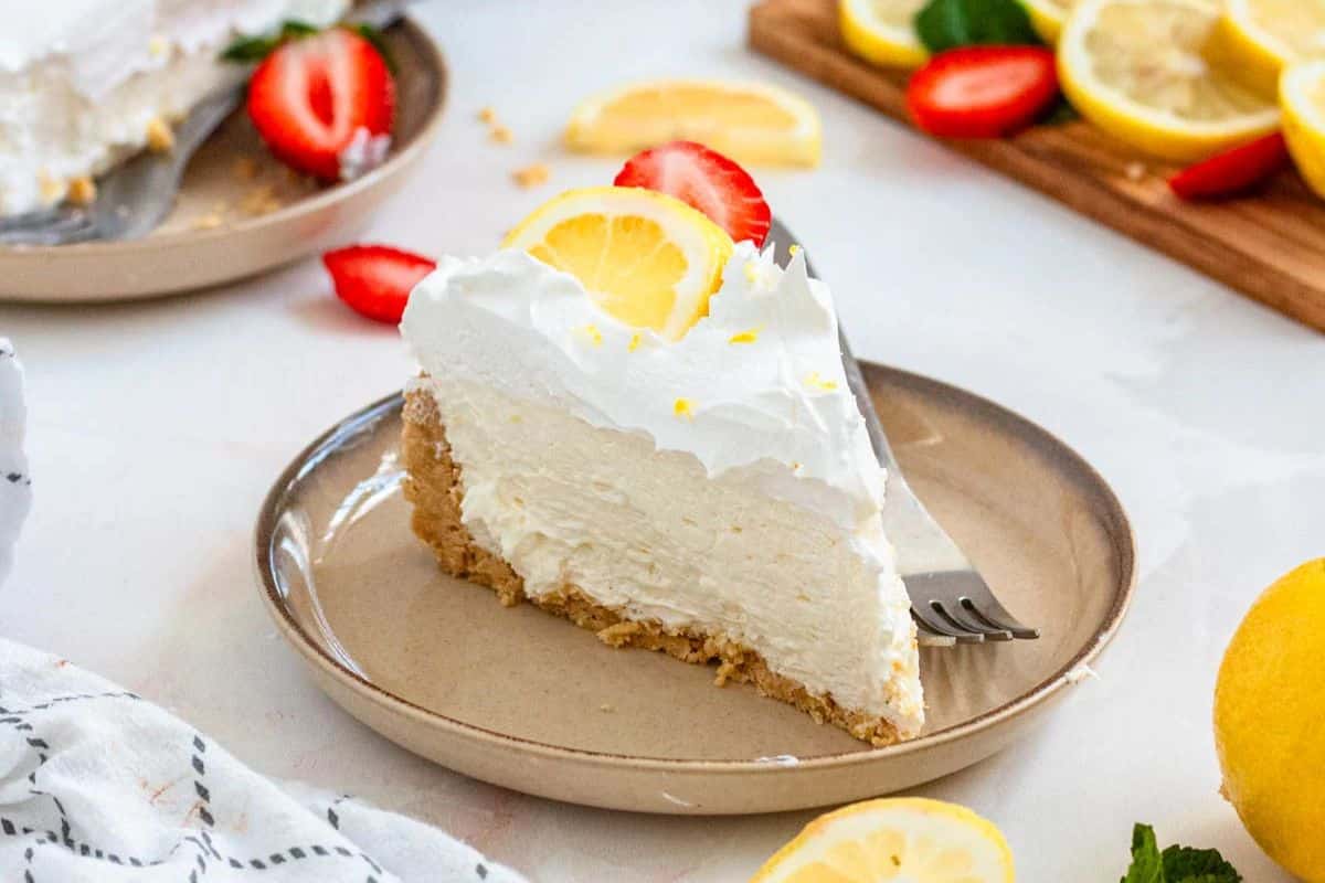 A slice of lemon cheesecake topped with whipped cream, a lemon slice, and a strawberry on a beige plate.