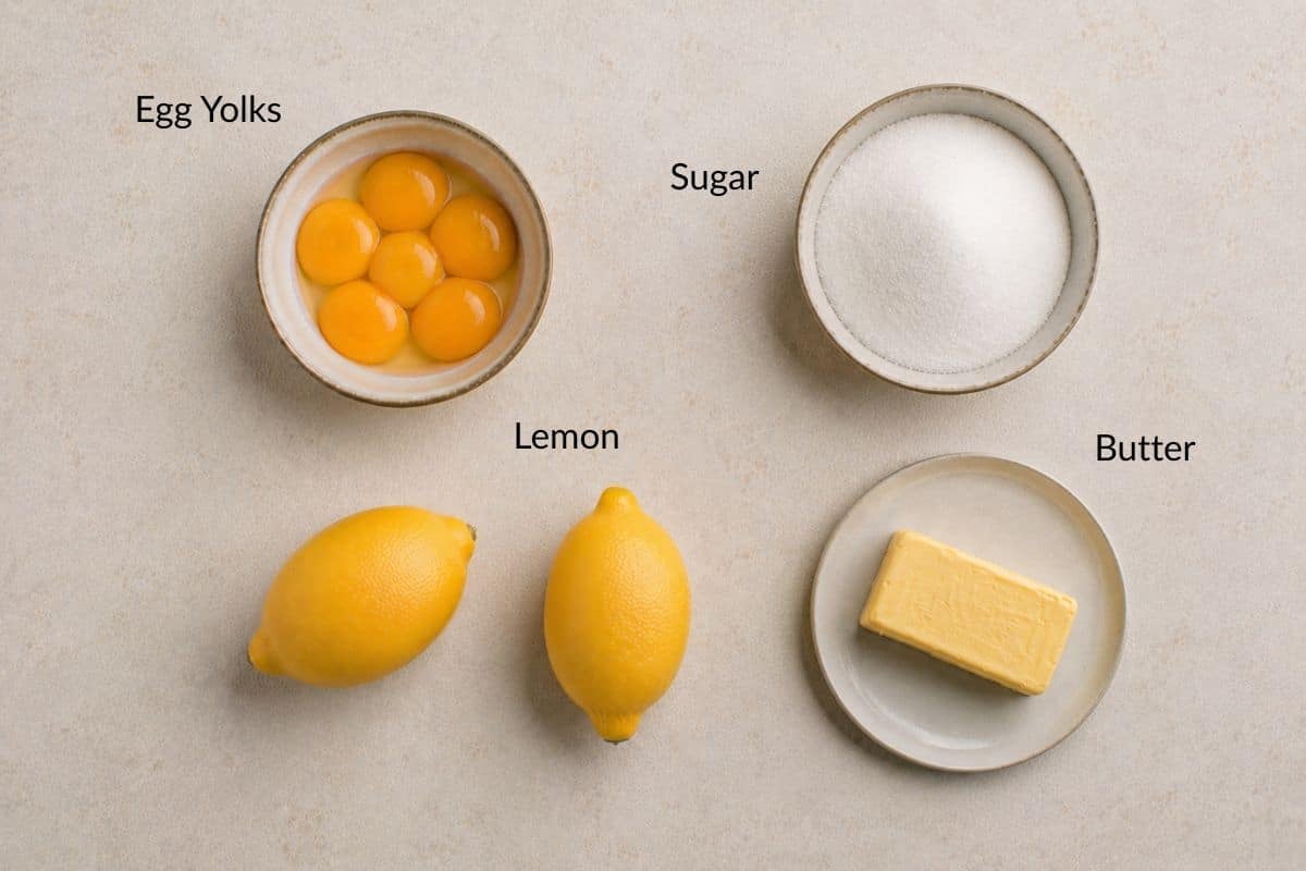 Four ingredients: a bowl of egg yolks, a bowl of sugar, two lemons, and a plate with a stick of butter.