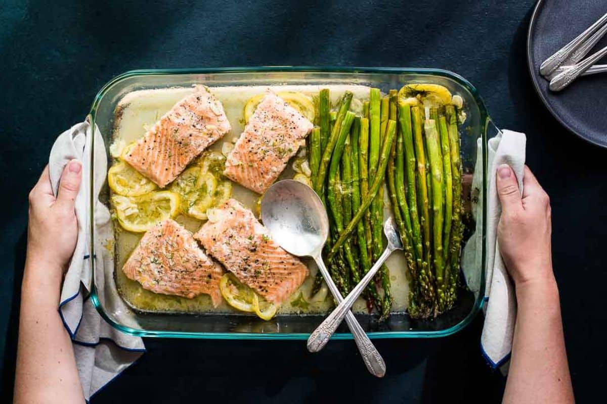 Person holding a baking dish with baked salmon on lemon slices and roasted asparagus, with serving utensils inside.