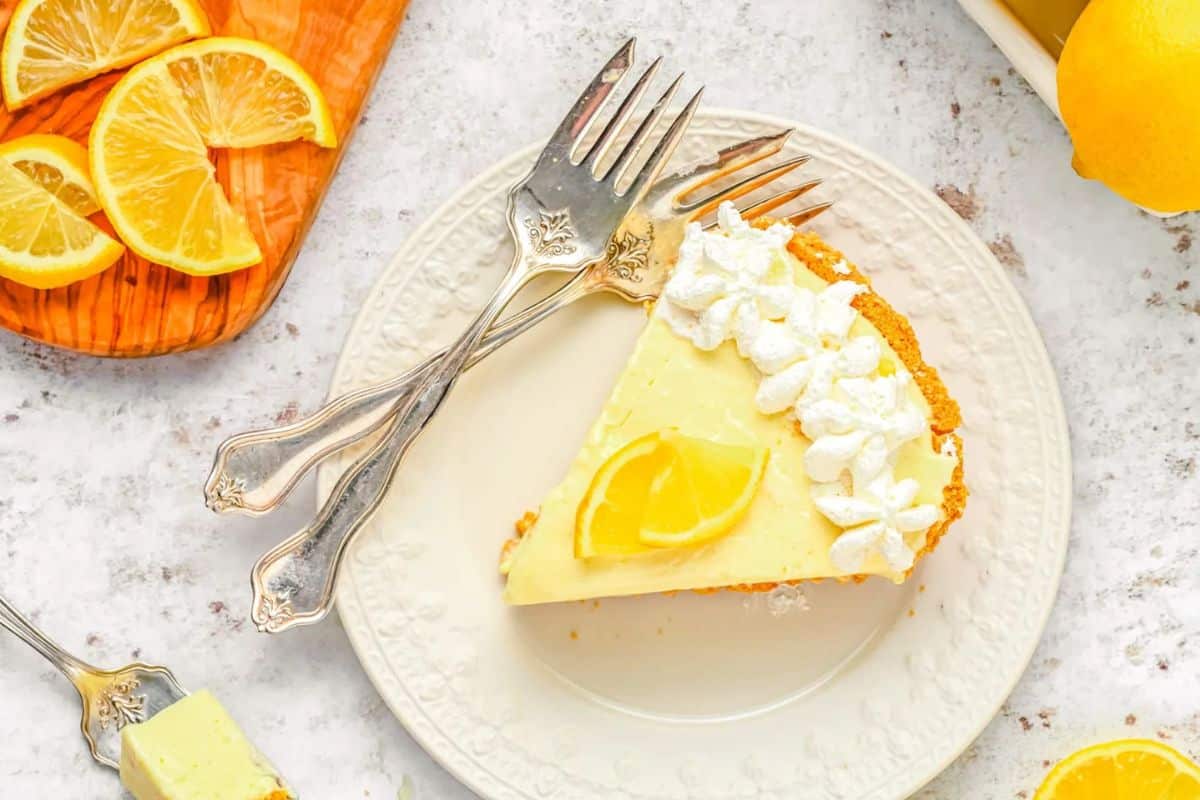 A slice of lemon pie with whipped cream and a lemon wedge on a plate, with two forks and lemon slices nearby.
