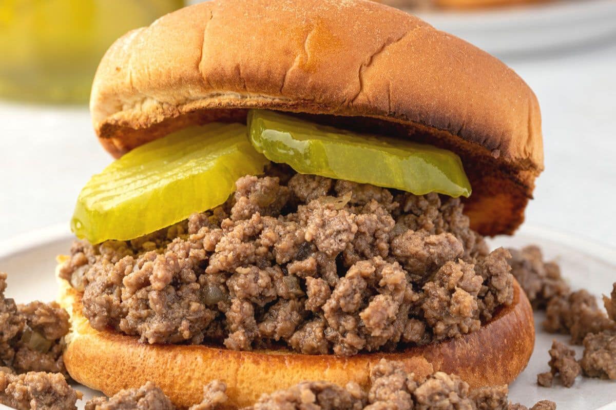 A sloppy joe sandwich with ground beef and two pickle slices on a toasted bun, with some meat spilled on the plate.