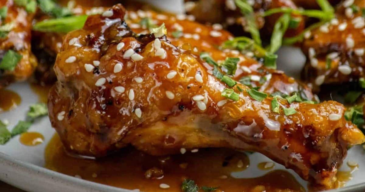 Close-up of glazed chicken drumstick topped with sesame seeds and chopped herbs on a white plate.