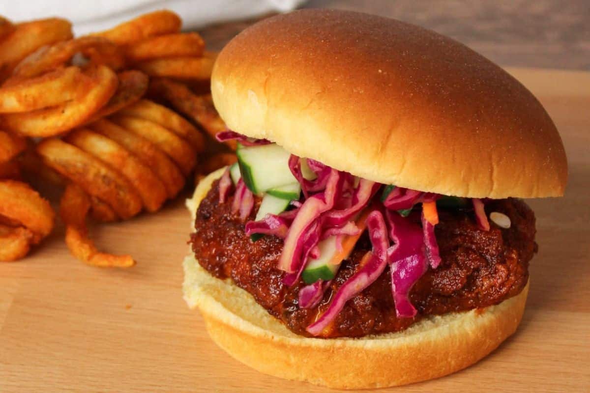 Fried chicken sandwich with coleslaw and cucumbers, served with a side of curly fries on a wooden surface.