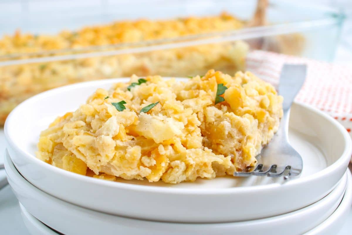 Two servings of cheesy hashbrown casserole on a white plate with a fork, topped with fresh herbs.
