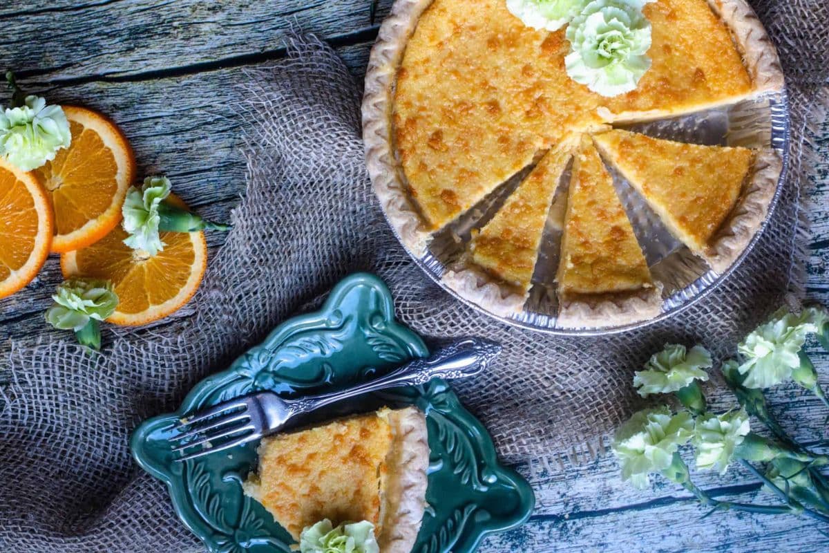 A sliced pie on burlap with orange slices and flowers, one slice served on a green plate with a fork.