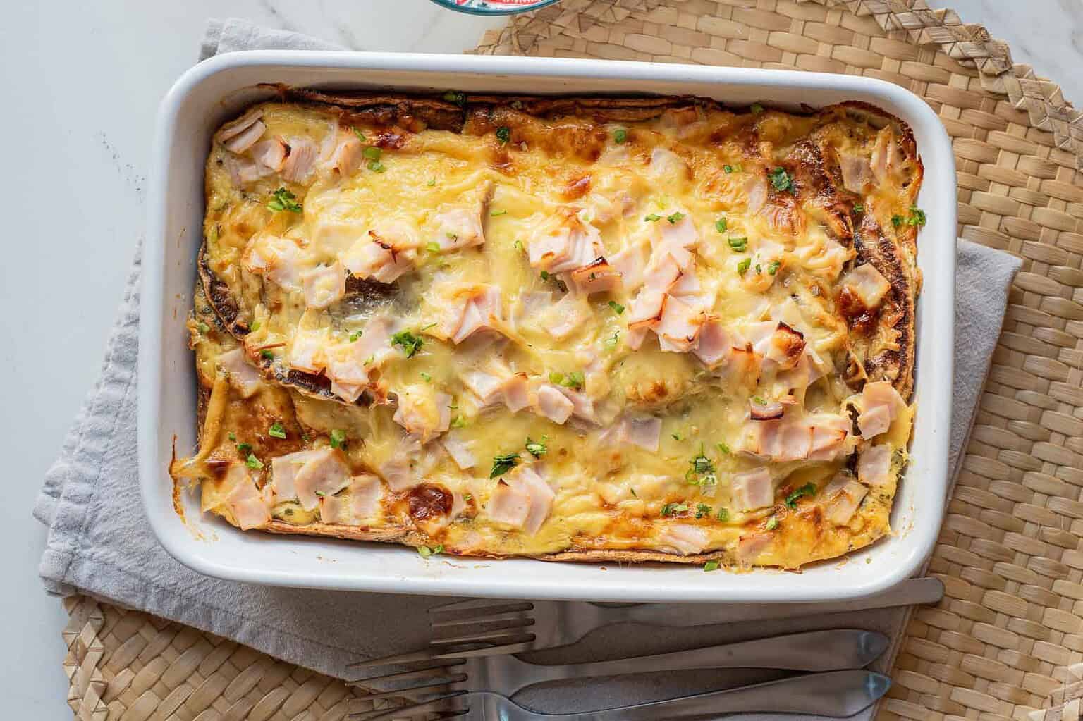 Baked casserole topped with melted cheese and diced ham in a white dish, on a woven placemat with utensils.