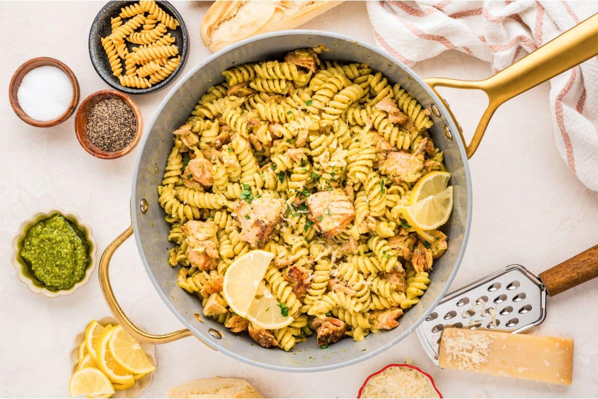 A skillet of pesto pasta with chicken, garnished with lemon slices, surrounded by cheese, seasonings, and pasta.