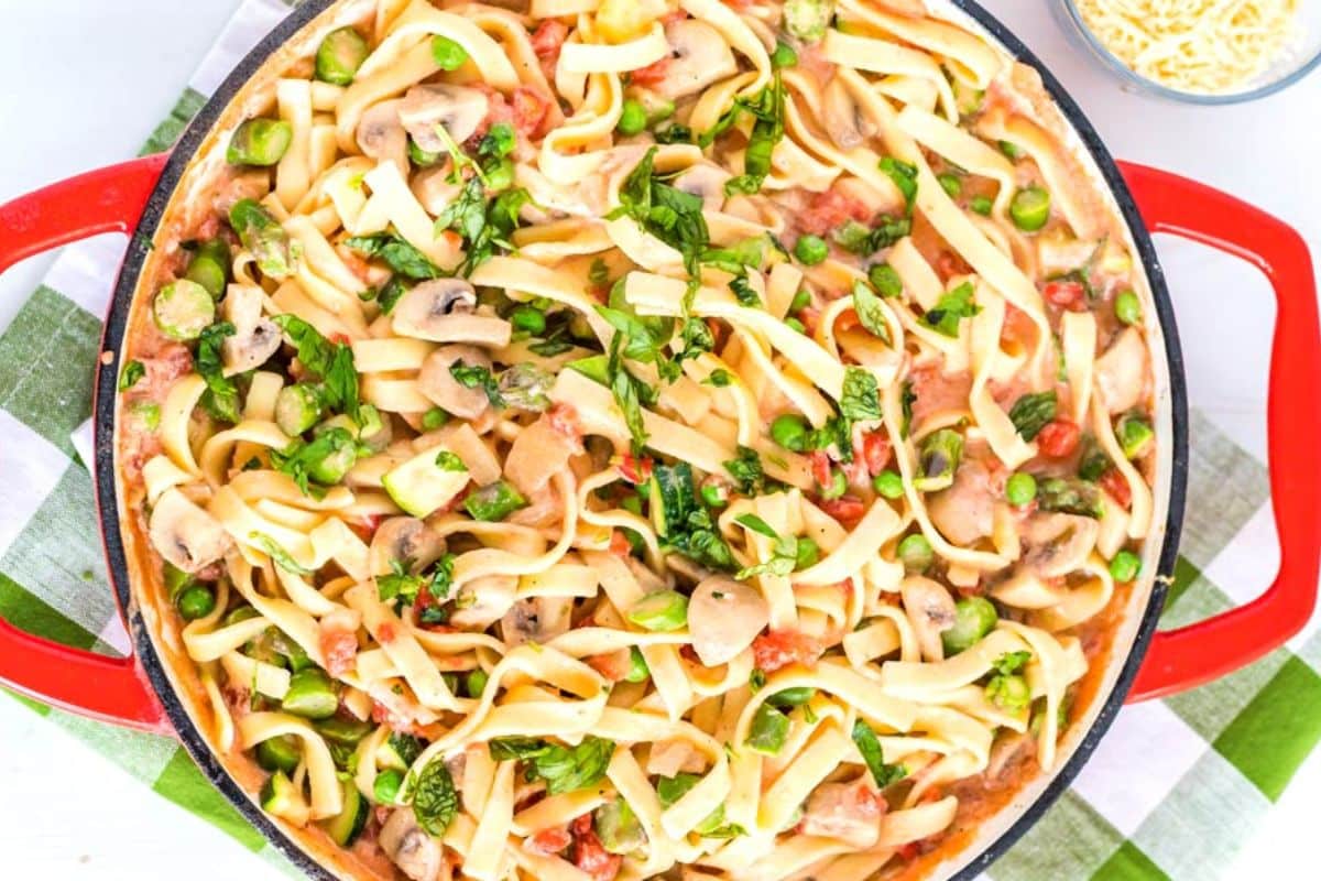 A red skillet filled with creamy pasta, mushrooms, peas, spinach, and tomatoes on a checkered cloth.