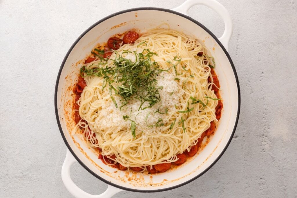 Cooked spaghetti in a white pot with tomato sauce, grated cheese, and chopped fresh basil on top.