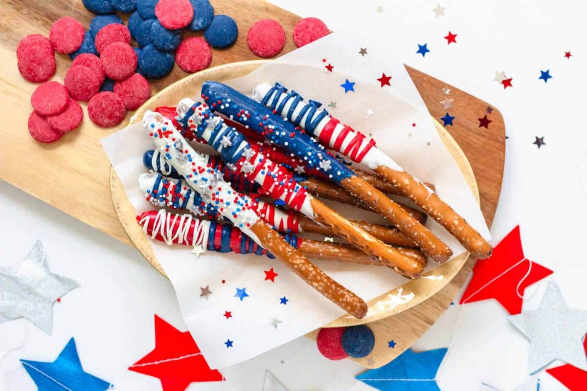 Pretzel rods dipped in red, white, and blue chocolate with star sprinkles on a plate, festive background.