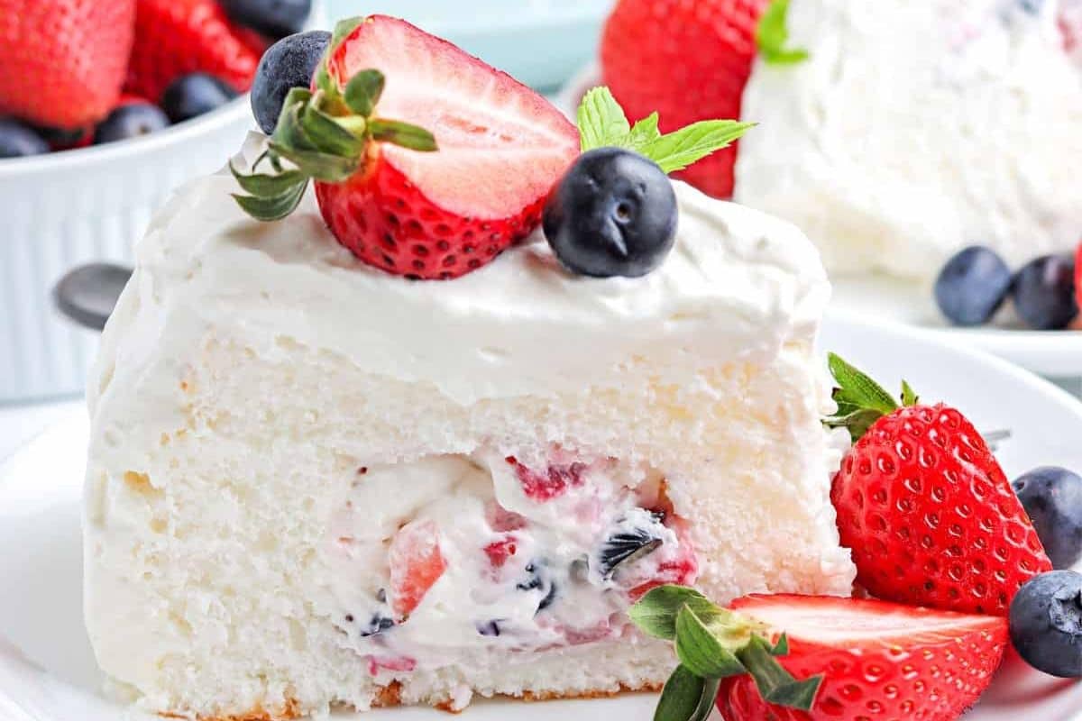 Slice of white cake with creamy filling, topped with whipped cream, strawberries, and blueberries.
