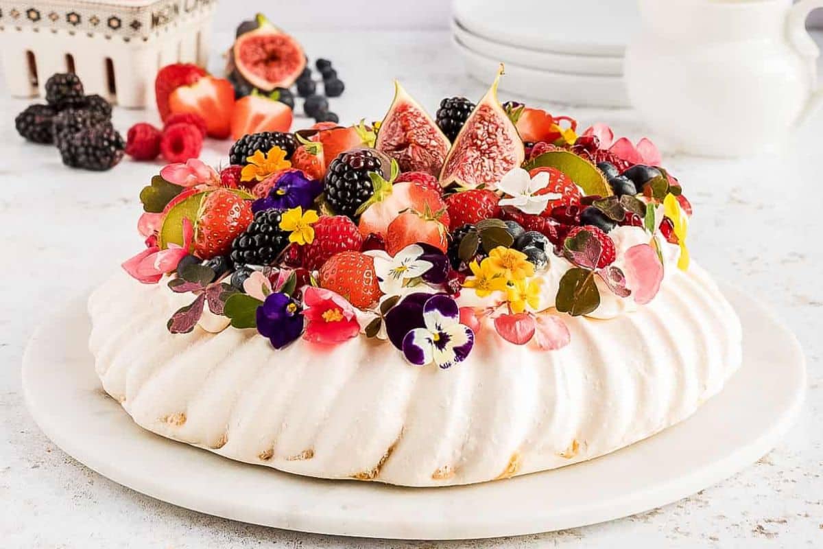 A pavlova topped with figs, berries, strawberries, and colorful edible flowers on a white plate.
