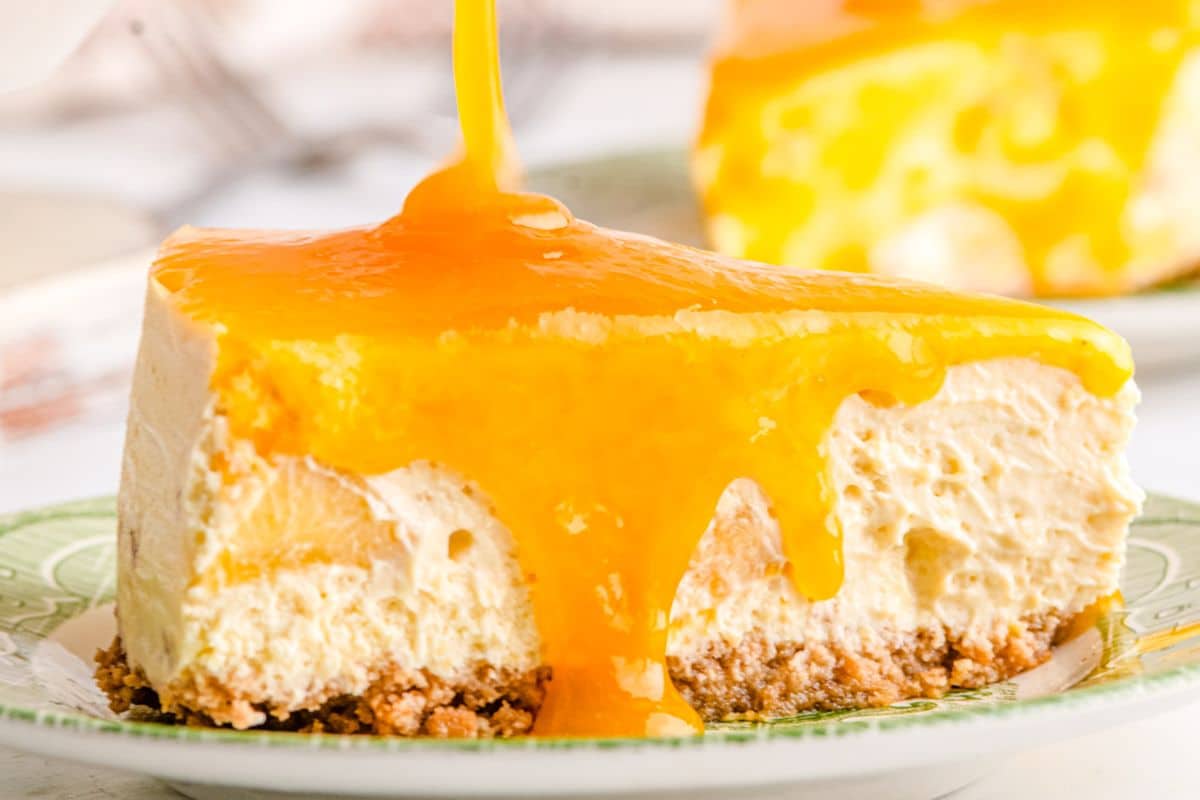 A slice of cheesecake with a graham cracker crust topped with thick, glossy orange sauce being poured over it.