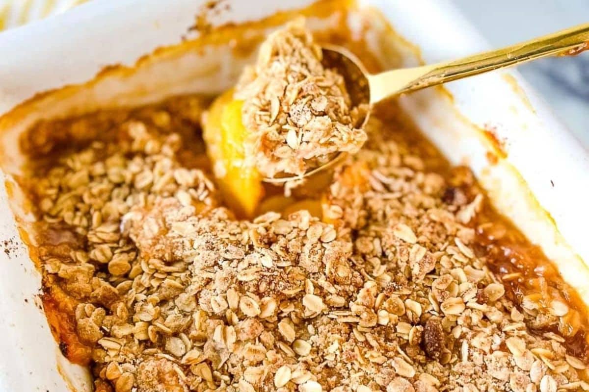 Golden peach crisp with oat crumble topping in a baking dish, a spoon scooping out a serving.