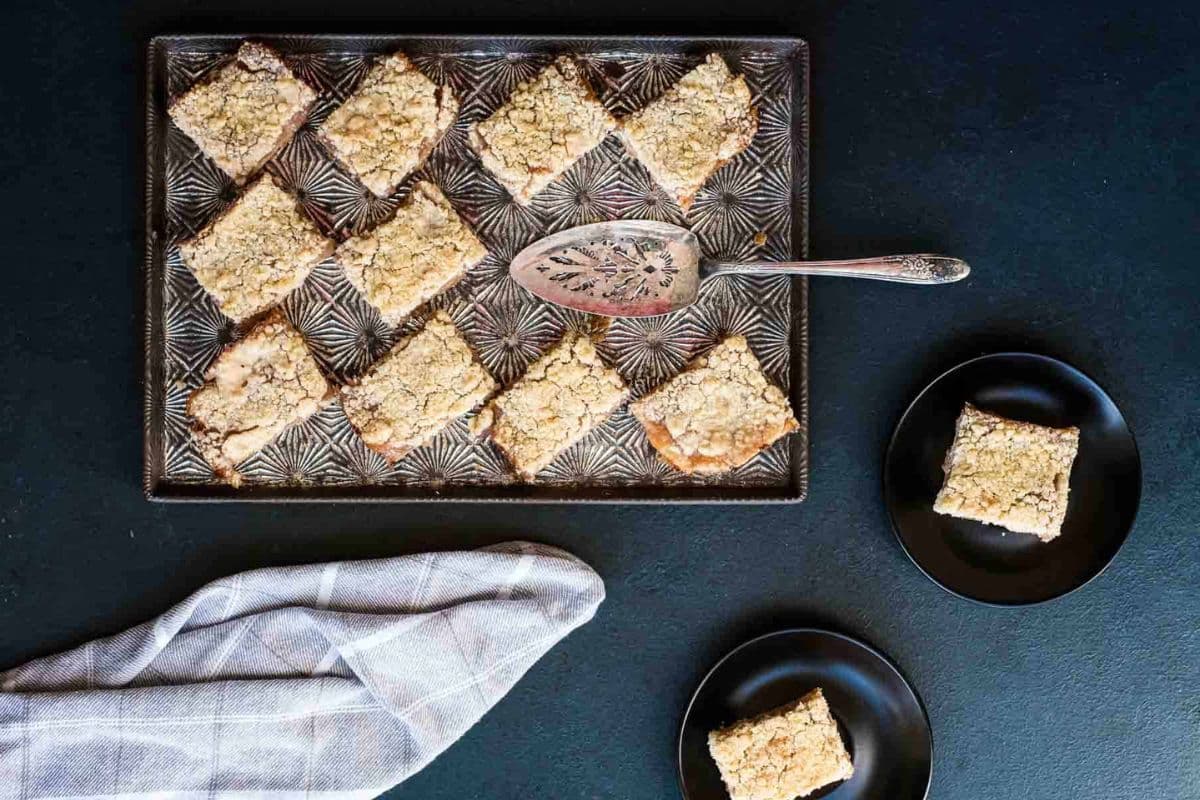 A tray of crumbly dessert bars with a serving utensil, two bars on black plates, and a checkered napkin nearby.