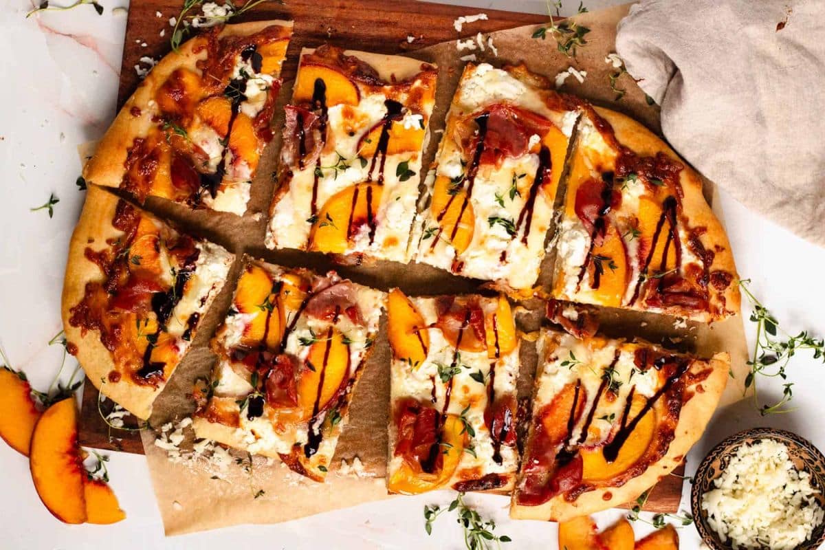 Sliced peach and prosciutto pizza with ricotta, balsamic glaze, and fresh herbs on parchment paper.