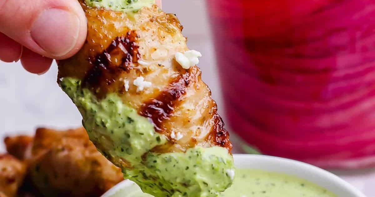 A grilled chicken wing dipped in creamy green sauce, held above a small bowl, with a pink drink in the background.