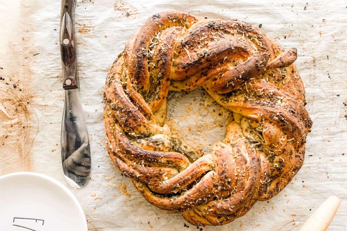 A twisted, golden-brown bread wreath topped with herbs and cheese on parchment paper next to a knife.