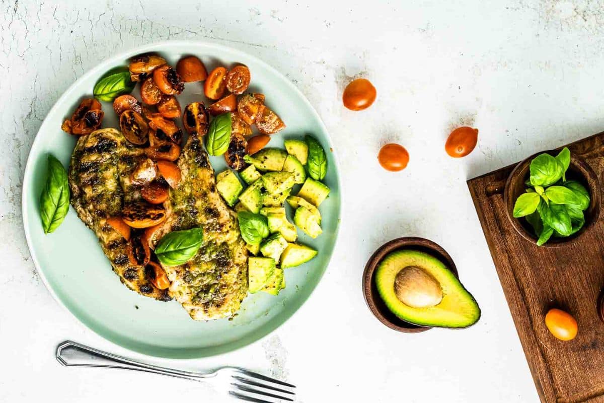 Grilled fish with tomatoes, avocado, and basil on a plate; fork and fresh ingredients on the side.