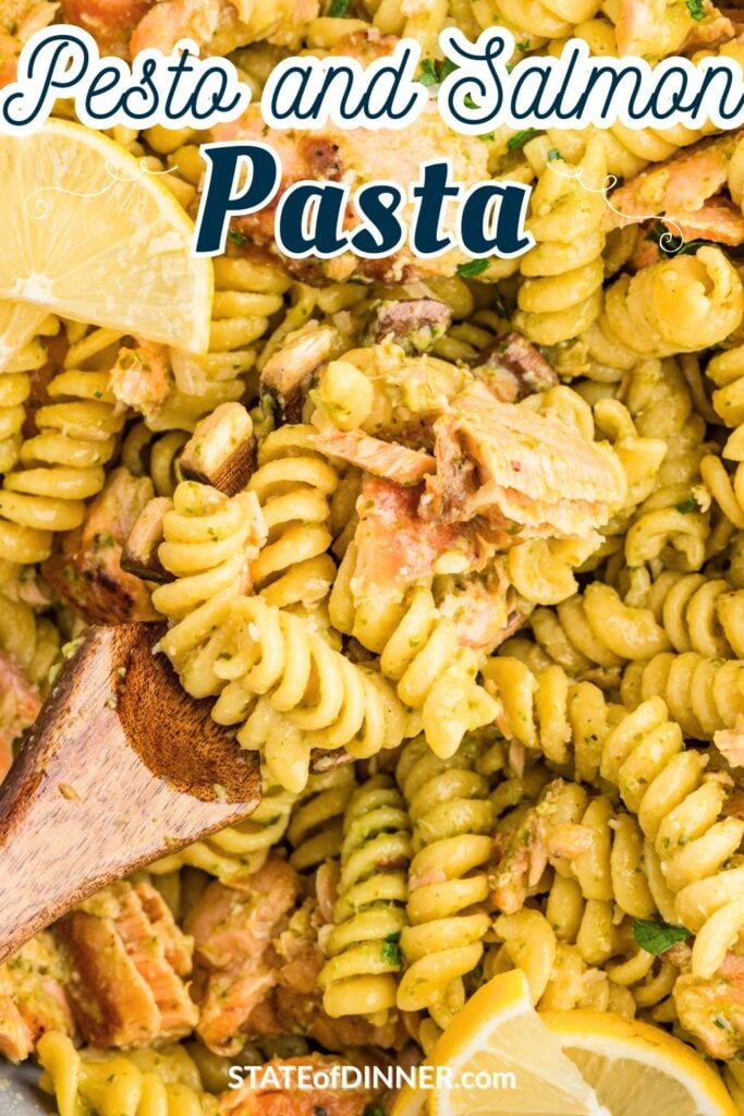 Rotini pasta with salmon chunks and pesto sauce, garnished with lemon wedges and served with a wooden spoon.