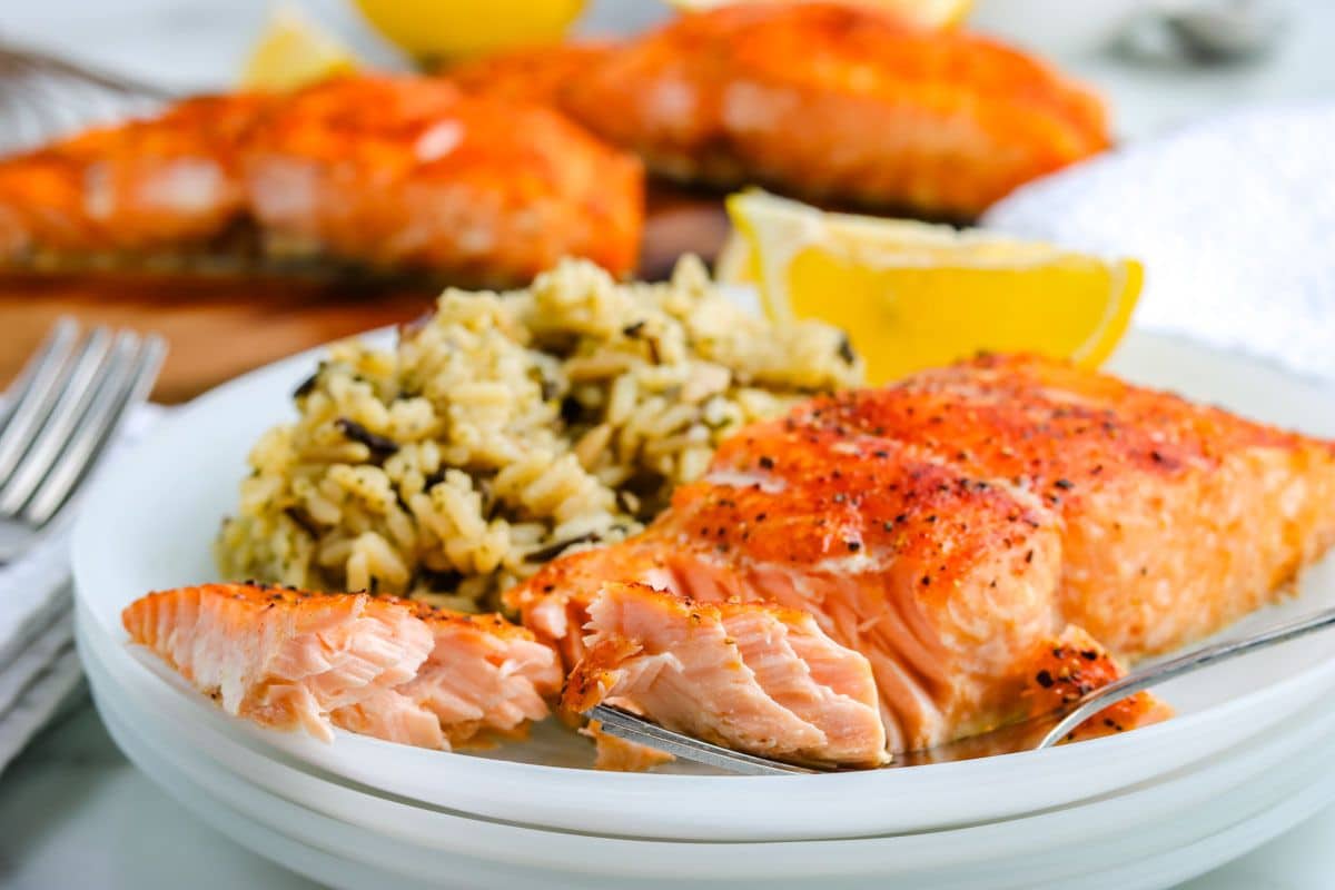 Grilled salmon fillet with wild rice and a lemon wedge on a white plate, with a forkful of salmon.