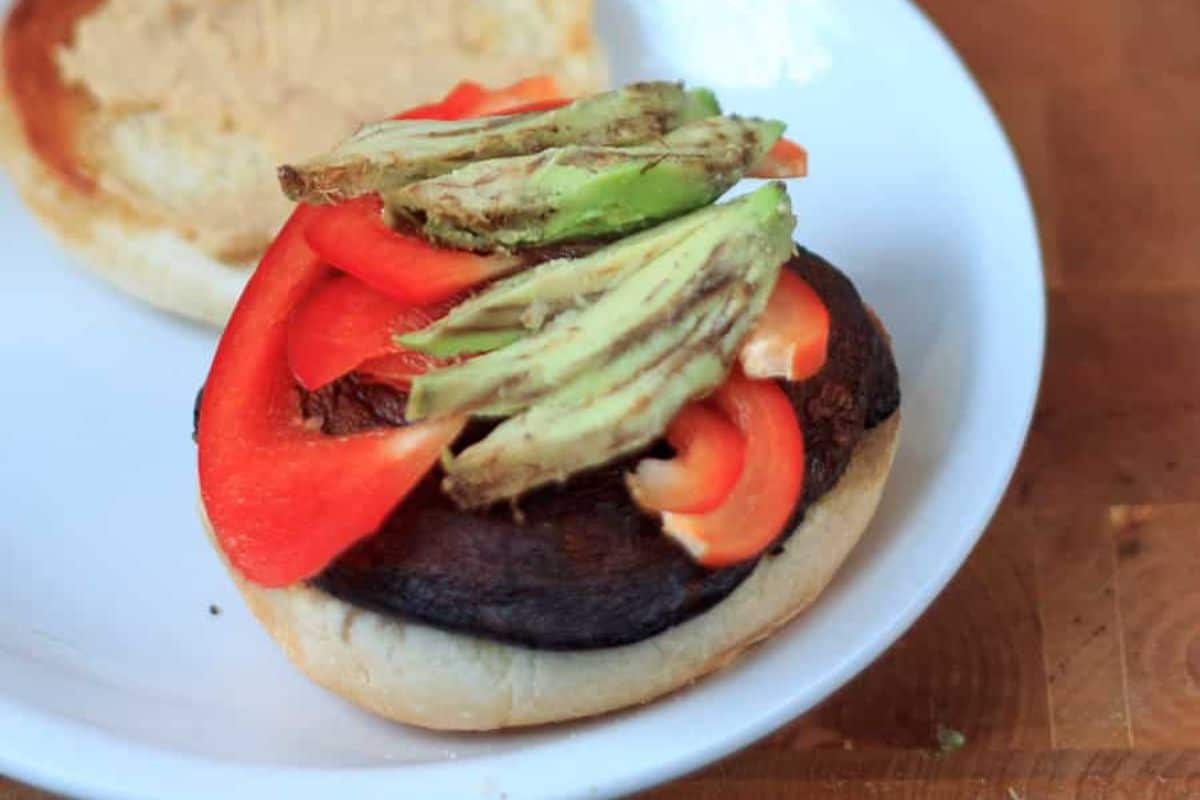 Open sandwich with a grilled portobello mushroom, red bell peppers, and avocado slices on a white plate.