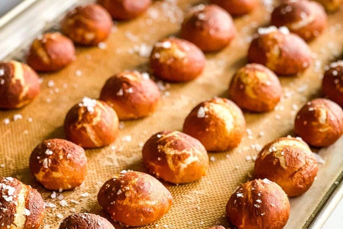 Rows of golden-brown pretzel bites sprinkled with coarse salt on a baking sheet lined with a silicone mat.