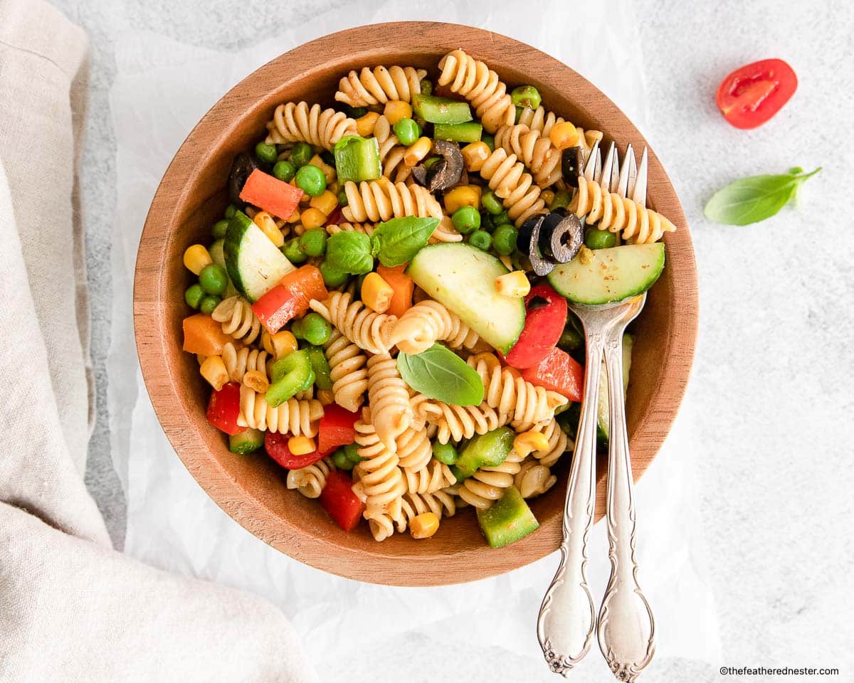 A wooden bowl of pasta salad with vegetables, garnished with basil, with two forks beside it.