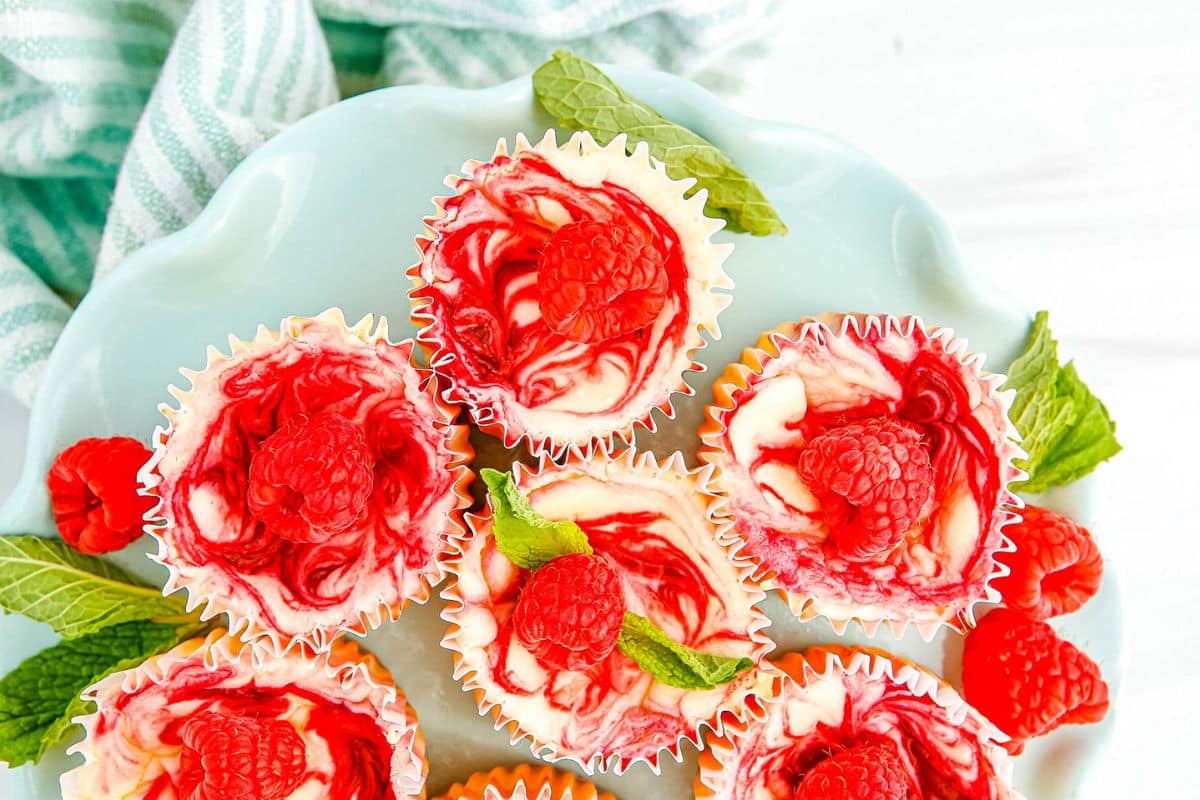 Mini cheesecakes with raspberry swirl, topped with fresh raspberries and mint, arranged on a light blue plate.