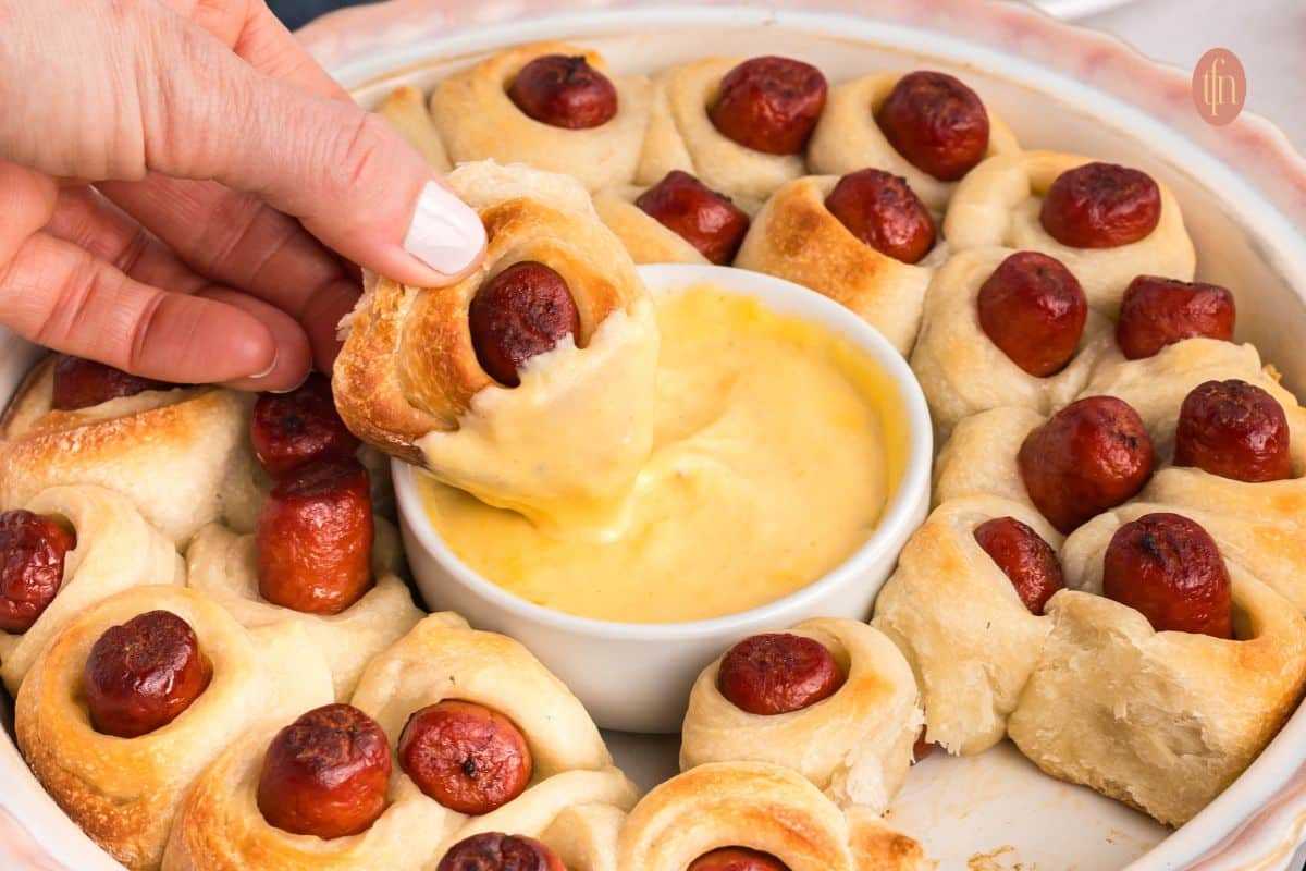 Hand dipping a pigs in a blanket roll into a bowl of cheese sauce, surrounded by more rolls on a platter.