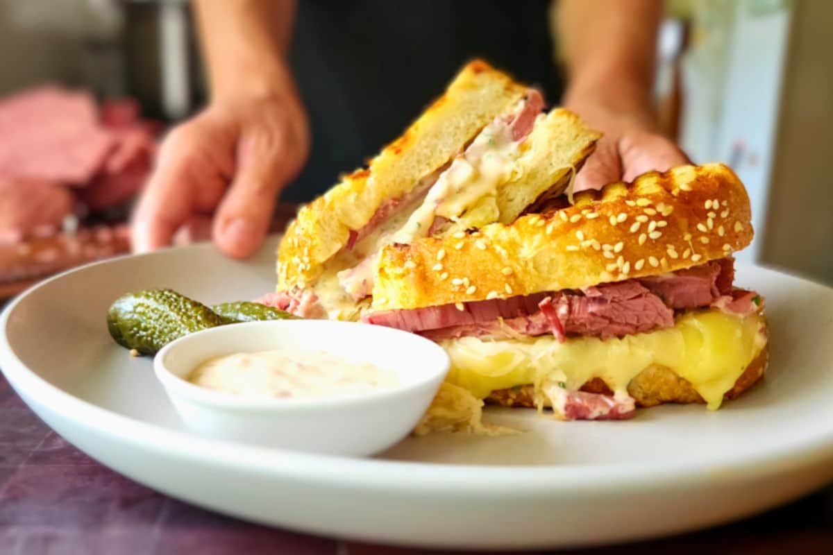 Close-up of a stacked sandwich with melted cheese, a side of pickles, and dipping sauce on a plate.