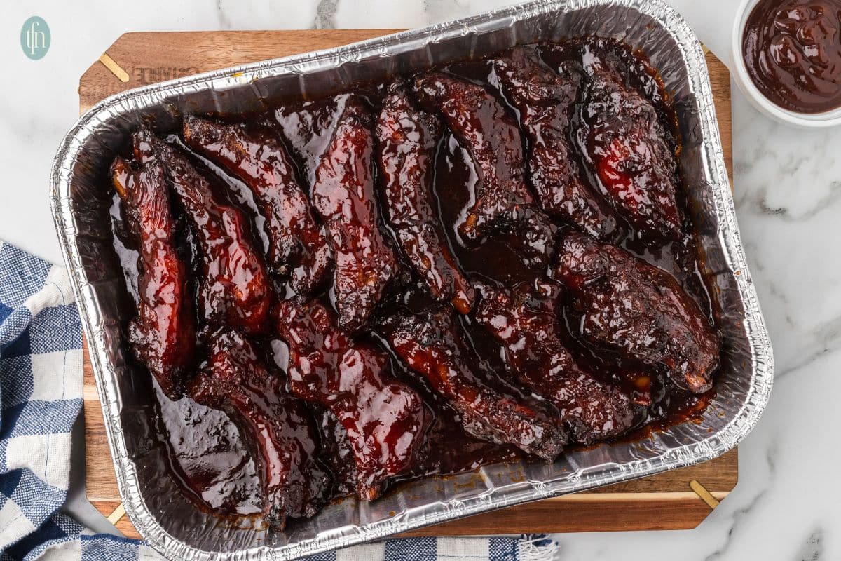 A foil tray filled with glazed barbecue ribs on a wooden board, with a blue and white towel beside it.