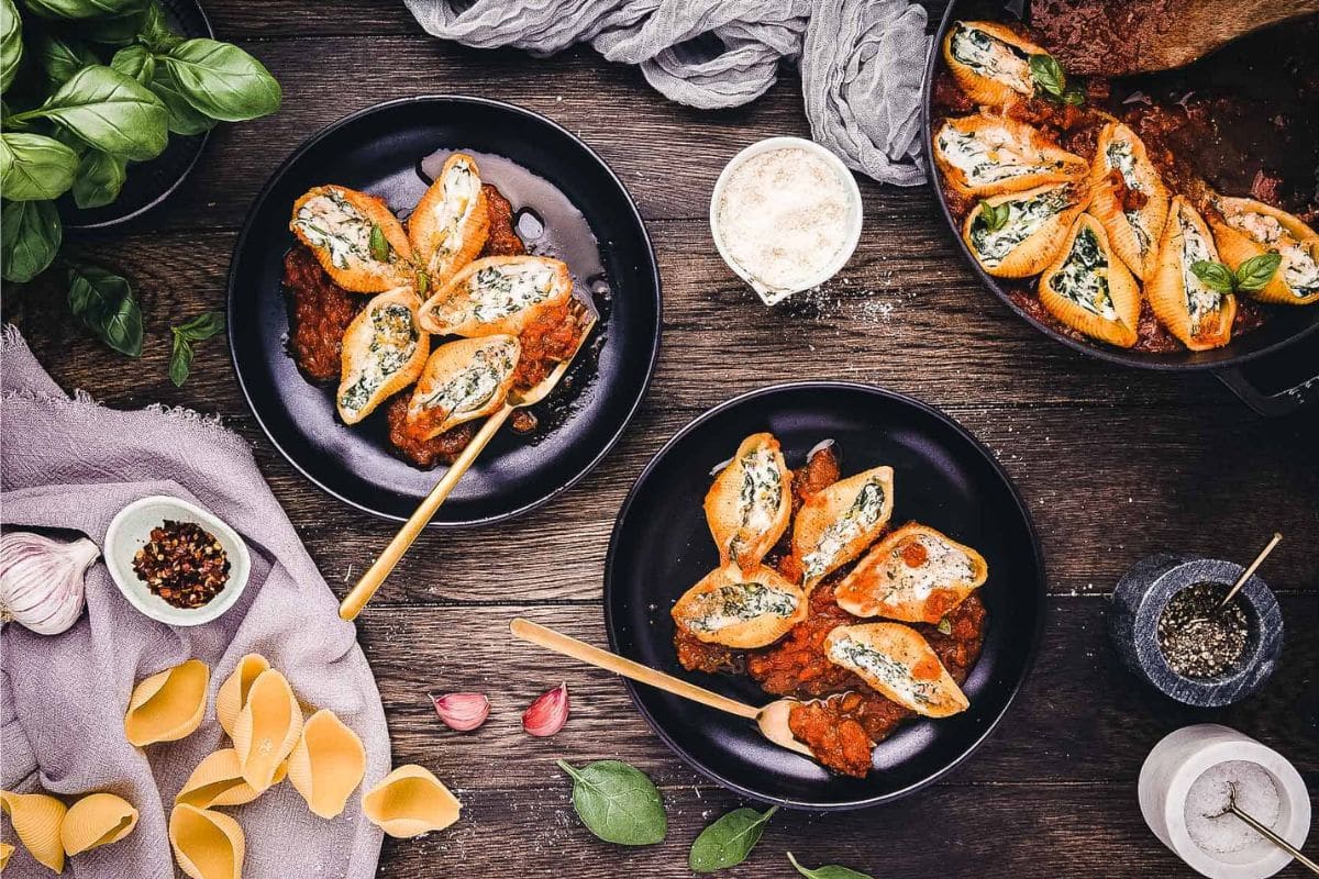 Three bowls of stuffed pasta shells with sauce, surrounded by herbs, spices, and uncooked pasta on a rustic table.