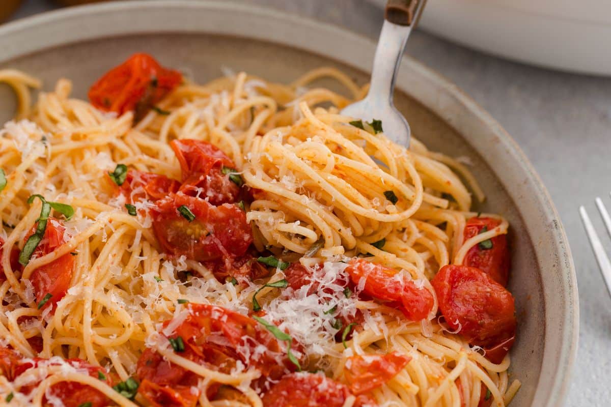 A bowl of spaghetti with cherry tomatoes, grated cheese, and fresh herbs, with a fork twirling pasta.