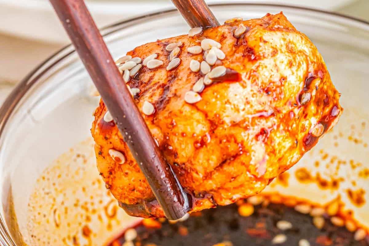 Chopsticks hold a spicy, sesame-topped tofu cube over a bowl of red chili sauce.
