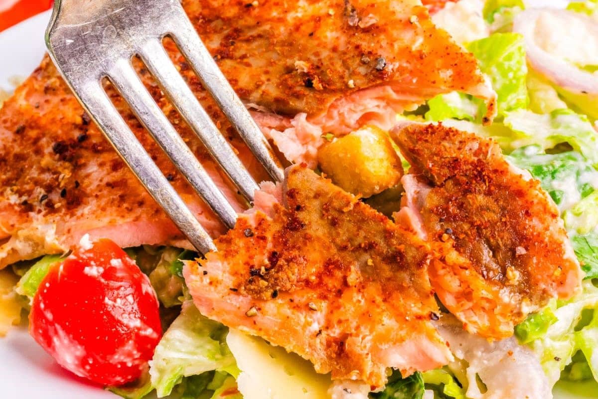 A fork holds a piece of seasoned, cooked salmon on a fresh salad with lettuce, tomato, and cheese.