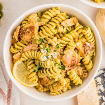A bowl of rotini pasta with pesto sauce, salmon chunks, grated cheese, parsley, and a lemon slice.