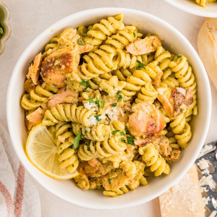 A bowl of rotini pasta with pesto sauce, salmon chunks, grated cheese, parsley, and a lemon slice.