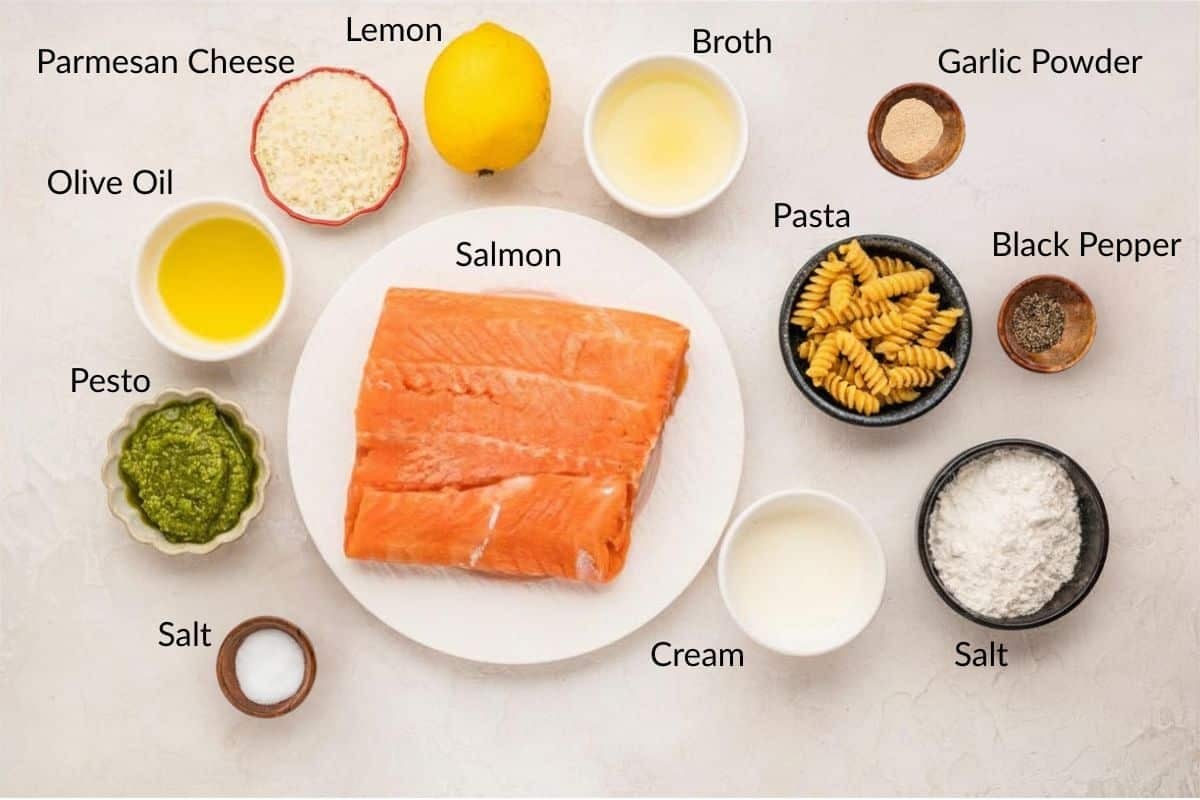 Overhead view of salmon, pasta, and various labeled ingredients in bowls, including olive oil, pesto, and cream.