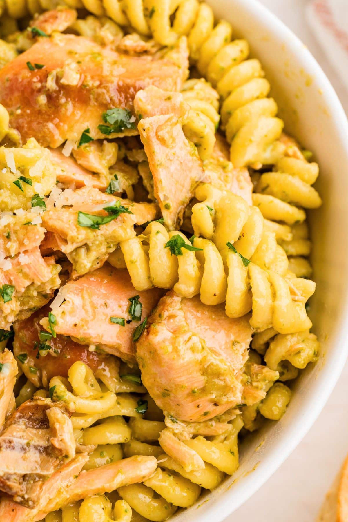 Close-up of rotini pasta with chunks of salmon and creamy sauce, garnished with fresh herbs in a white bowl.
