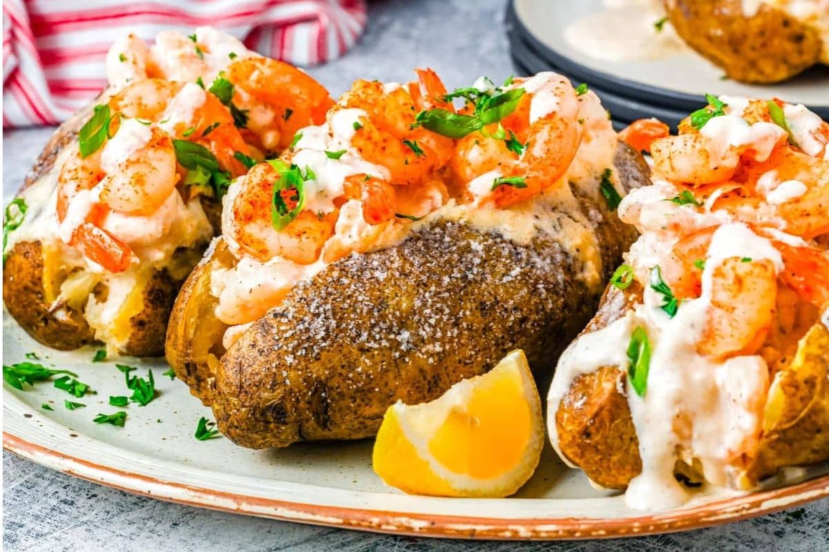 Three baked potatoes topped with creamy shrimp and herbs sit on a plate with a lemon wedge.